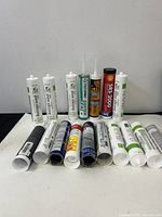 Photo showing 16 new adhesive silicone tubes of various brands and types, arranged in two rows on a flat surface against a plain wall.