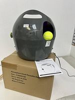 Gray automatic pet ball launcher sitting on a box with an included tennis ball placed in the ball chamber. Instruction manual and power cord visible.