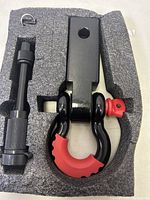 Close-up photo of the shackle hitch set in foam packaging showing the U-shaped shackle, red protective cover, hitch pin, and securing clip.