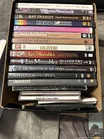 Box of assorted music and musical DVDs stacked with spines showing titles including Riverdance, Ray Charles, Les Miserables, Phantom of the Opera, Amadeus and others.