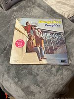 Front cover of Stampeders Carryin' On vinyl LP showing band members sitting outdoors by a bridge, with album title and sticker for hit single Wild Eyes.