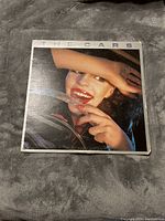 Front cover of The Cars self-titled vinyl LP showing a woman smiling behind a car steering wheel with the band name at the top.