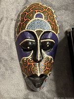 Front-facing photo of the wooden tribal mask showing detailed dot art in multiple colors including purple, light blue, orange, white, and black.