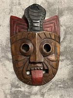 Front view of a hand-carved wooden mask with a cobra figure on the forehead, deep facial carvings, round eye holes, protruding red tongue, and painted details on the serpent.