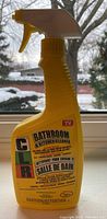 Full front view of yellow CLR Bathroom & Kitchen Cleaner spray bottle on window sill with background outdoor scene.