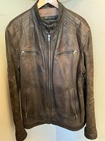 Front view of brown leather men's jacket, showing zippered chest and side pockets, full zipper closure, and buckle strap at collar.