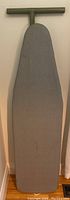 Ironing board standing upright against the wall with grey padded cover visible, showing the T-shaped handle at the top and tapered shape of the board.