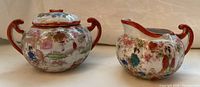Photo showing front view of Japanese ceramic sugar bowl and creamer with floral and garden patterns, reddish-brown handles, and gold detail. Sugar bowl lid handle is broken.