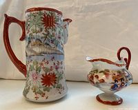 Side views of one tall and one short Japanese porcelain serving jug with detailed hand-painted decorations, gold accents, and red handles.