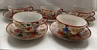 Photo showing five hand painted porcelain Japanese tea cups each paired with decorated saucers. Cup and saucer designs feature colorful traditional Japanese figures and floral patterns. Pieces require cleaning.