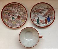 Top view of two small painted porcelain plates and one tea cup showing hand-painted traditional Japanese designs with geisha, floral patterns, and a red edge rim.