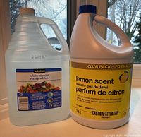 Photo showing two large plastic jugs: one labeled lemon scent bleach 3.6L with yellow label, one labeled white vinegar 2.5L with blue and white label. Both unopened.