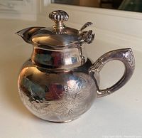 Side view of the rounded personal teapot showing floral engraving, scalloped spout rim, and curved handle with ornate detailing.
