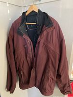 Front view of burgundy outer shell winter coat on wooden hanger showing hood and zipper closure