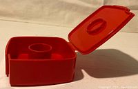 Red square tape dispenser with hinged lid open, showing interior spool and tape groove.