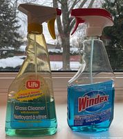 Two spray bottles of glass cleaner placed on a window sill with a snowy outdoor view. The left bottle is Life brand, Glass Cleaner with Ammonia, partially used with yellow spray top. The right bottle is Windex Original, partially used with a red spray top.
