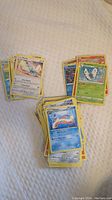 Multiple stacks of modern Pokémon trading cards on a textured white surface, featuring recognizable Pokémon including a Vaporeon card in the foreground and a Butterfree card in another stack.