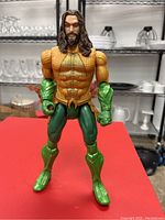 Full body shot of Aquaman figure standing on red surface showing orange and green suit with muscular build.