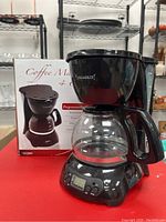 Front view of the black programmable coffee maker next to its original box, showing glass carafe and overall shape and size.