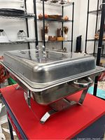 Photo of rectangular stainless steel deep pan chafer dish with lid and handles on a red surface, showing its profile and stand below.