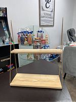 View of the two-tier wooden hanging bookshelf with gold metal supports on either side, placed on a table with a background of cosmetics on glass shelves.