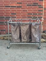 Full view of metal frame laundry sorter with three fabric bags on wheels against a brick wall.