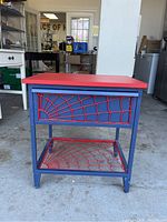 Front view of the metal cabinet showing the red spider web design on the drawer front and the lower shelf, blue frame, and red top surface.