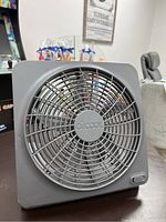 Front view of the grey O2 Cool battery powered fan showing the circular grille and fan blades beneath, with the brand name embossed at center.