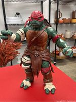 Front view of the TMNT figurine showing green turtle with red eye mask, brown chest armor, and bandage wrappings on limbs.