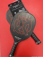 Two V2 Recruit Onix pickleball paddles lying on red surface, showing front face with black and red logo.
