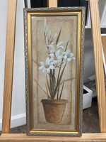 Full view of tall narrow framed wall art with white flowers in a brown pot on beige background.