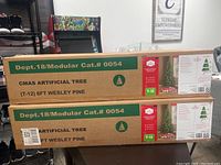 Boxes of two Wesley Pine 6-foot artificial Christmas trees, marked as non-lit with plastic tree stands. Shows full packaging details including size, branch tips, and product code T-12.