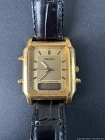 Front view of watch showing gold-tone rectangular case, champagne dial, analog hands and digital LCD at 6 o'clock, black leather strap partially visible.