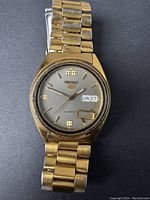 Front view of vintage Seiko automatic men's watch showing gold-tone metal case and bracelet, grey dial with Seiko 5 logo, day-date display, and detached date window frame.
