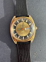 Front view of vintage Caravelle automatic wristwatch with black and gold dial, day-date window, and black textured leather strap. Shows patina and wear on case.