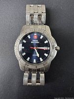 Front view of Swiss Military watch showing black dial with white markers, red second hand, date window, and stainless steel case and bracelet.
