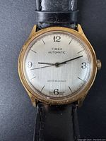 Front view showing gold-tone case, silver dial with black numerals, and black leather strap.