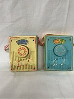 Front view of two vintage Fisher Price music box pocket radios from 1968, one cream colored and one blue, showing turn knobs and musical decorations.