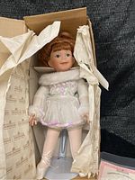 Porcelain Ashton Drake doll in white ballerina outfit in original box, doll standing on a display stand with ribbons on ballet shoes.