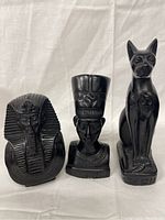 Front view of three black Egyptian figurines on white background: pharaoh bust, queen/goddess bust with tall headdress, seated cat statue.
