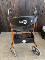 Front view of foldable orange rollator walker with black seat and 'hugo' on backrest.