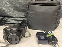 Front view of Panasonic Lumix DMC-FZ20 digital camera with lens cap, wrist strap, charger, power cable, and black soft carrying case.