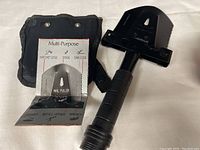 Photo showing multi-purpose hand tool next to its black carrying pouch. The tool head displays labels for hatchet edge, spade, saw edge, nail puller, hammer, bottle opener, and wrench.