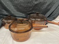 Photo showing three amber-brown Pyrex glass pots with lids and long handles of varying sizes arranged on a white surface with a dark fabric background.