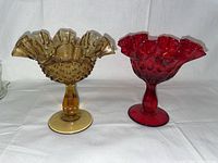 Two vintage glass compote pedestal dishes side by side, amber hobnail and ruby red thumb print, showing overall shape and colors
