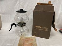 Photo showing the vacuum coffee maker assembled with the handle, glass chambers, black lid, and original box behind it. Instruction sheet is placed in front.