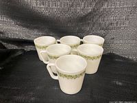 Front view of six white opaque Pyrex mugs with green flower pattern and D handles arranged in two rows on dark textured fabric background.