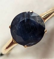 Close-up image of a 14K gold ring with a deep blue round sapphire held by four prongs, showing gemstone facets and natural inclusions.