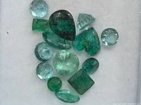 Multiple emerald gemstones of different cuts and shades displayed on a white background.