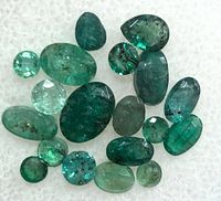 Close-up photo showing 21 natural emerald gemstones of various shapes and sizes on a white background.